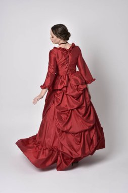 Kırmızı ipek Victoria elbisesi giyen esmer bir kızın tam boy portresi. Beyaz bir stüdyonun arka planında kameraya karşı durmak..