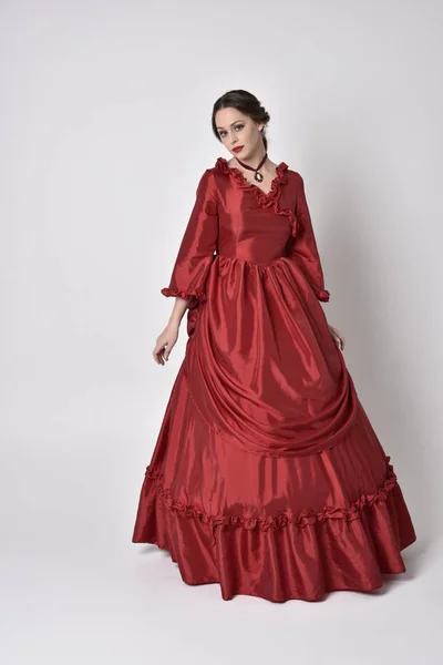 full length portrait of a brunette girl wearing a red silk victorian gown. Standing pose on a white studio background.