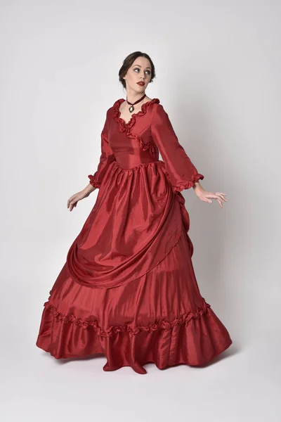 full length portrait of a brunette girl wearing a red silk victorian gown. Standing pose on a white studio background.