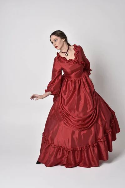 full length portrait of a brunette girl wearing a red silk victorian gown. Standing pose on a white studio background.