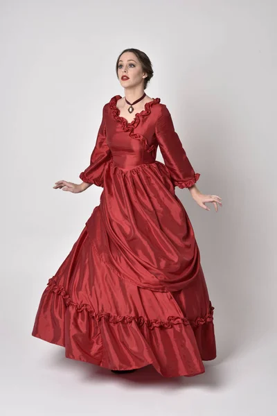 full length portrait of a brunette girl wearing a red silk victorian gown. Standing pose on a white studio background.