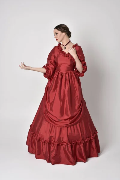 full length portrait of a brunette girl wearing a red silk victorian gown. Standing pose on a white studio background.