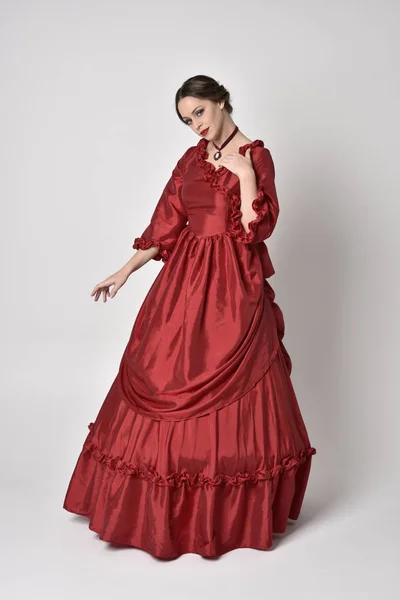 full length portrait of a brunette girl wearing a red silk victorian gown. Standing pose on a white studio background.