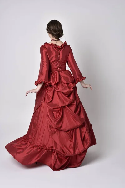 full length portrait of a brunette girl wearing a red silk victorian gown. Standing with back to the camera on a white studio background.