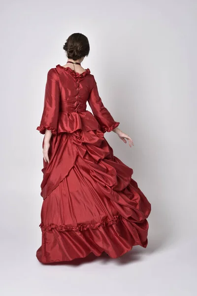 full length portrait of a brunette girl wearing a red silk victorian gown. Standing with back to the camera on a white studio background.