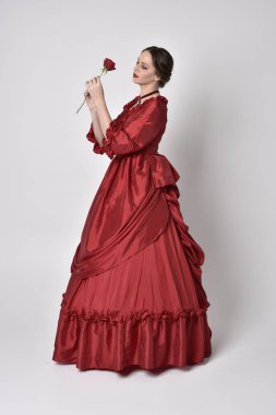  Kırmızı ipek Victoria elbisesi giyen esmer bir kızın portresi. Beyaz bir stüdyonun arka planında kameraya karşı durmak..