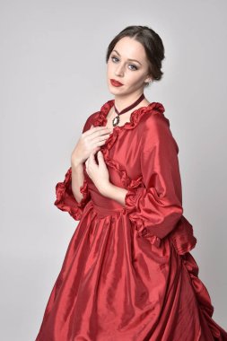  Kırmızı ipek Victoria elbisesi giyen esmer bir kızın portresi. Beyaz bir stüdyonun arka planında kameraya karşı durmak..