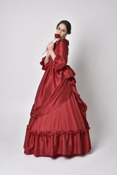 portrait of a brunette girl wearing a red silk victorian gown. Standing with back to the camera on a white studio background.