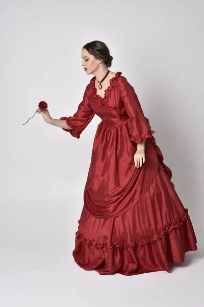 portrait of a brunette girl wearing a red silk victorian gown. Standing with back to the camera on a white studio background.