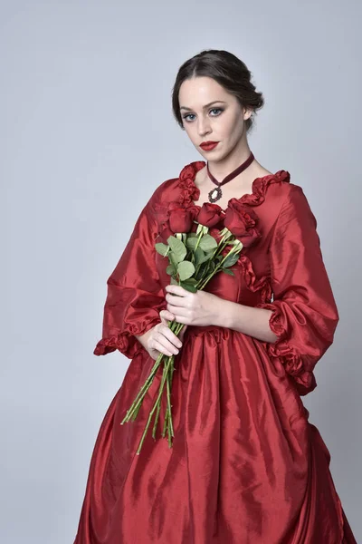 portrait of a brunette girl wearing a red silk victorian gown. Standing with back to the camera on a white studio background.