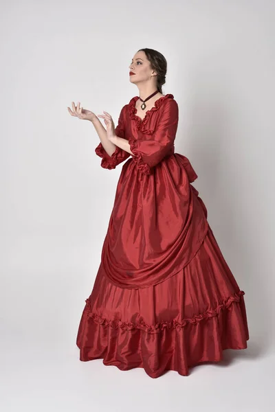 full length portrait of a brunette girl wearing a red silk victorian gown. Standing with back to the camera on a white studio background.