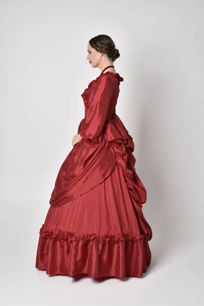 full length portrait of a brunette girl wearing a red silk victorian gown. Standing with back to the camera on a white studio background.