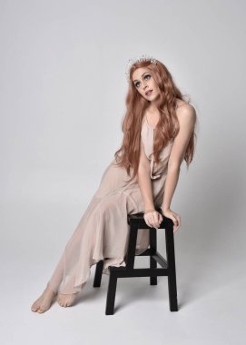 Çıplak, akıcı bir elbise ve kristal taç giyen bir kızın tam boy portresi. Gri bir stüdyonun arka planında bir sandalyede oturmak.