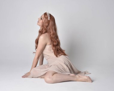 full length portrait of a pretty, fairy girl wearing a nude flowy dress and crystal crown. Seated pose against a grey studio background.