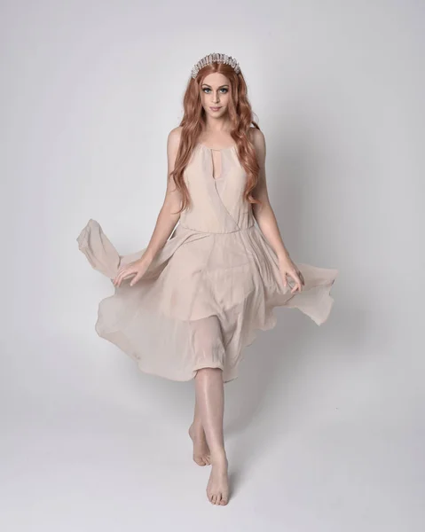 full length portrait of a pretty, fairy girl wearing a nude flowy dress and crystal crown. Standing and dancing pose against a grey studio background.