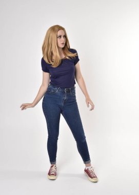 Sıradan mavi gömlek, kot pantolon ve spor ayakkabı giyen sarışın güzel bir kızın tam boy portresi. Stüdyo arka planında poz vermek.