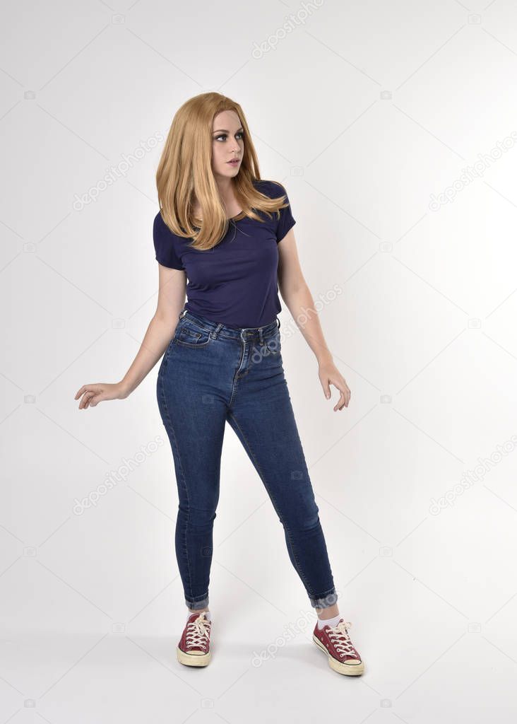Full length portrait of a pretty blonde girl wearing casual blue shirt, denim jeans and sneakers. Standing pose on a studio background.