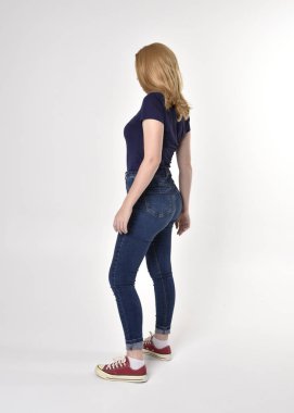 Sıradan mavi gömlek, kot pantolon ve spor ayakkabı giyen sarışın güzel bir kızın tam boy portresi. Poz vermek, kameradan uzaklaşmak, stüdyo arka planında....