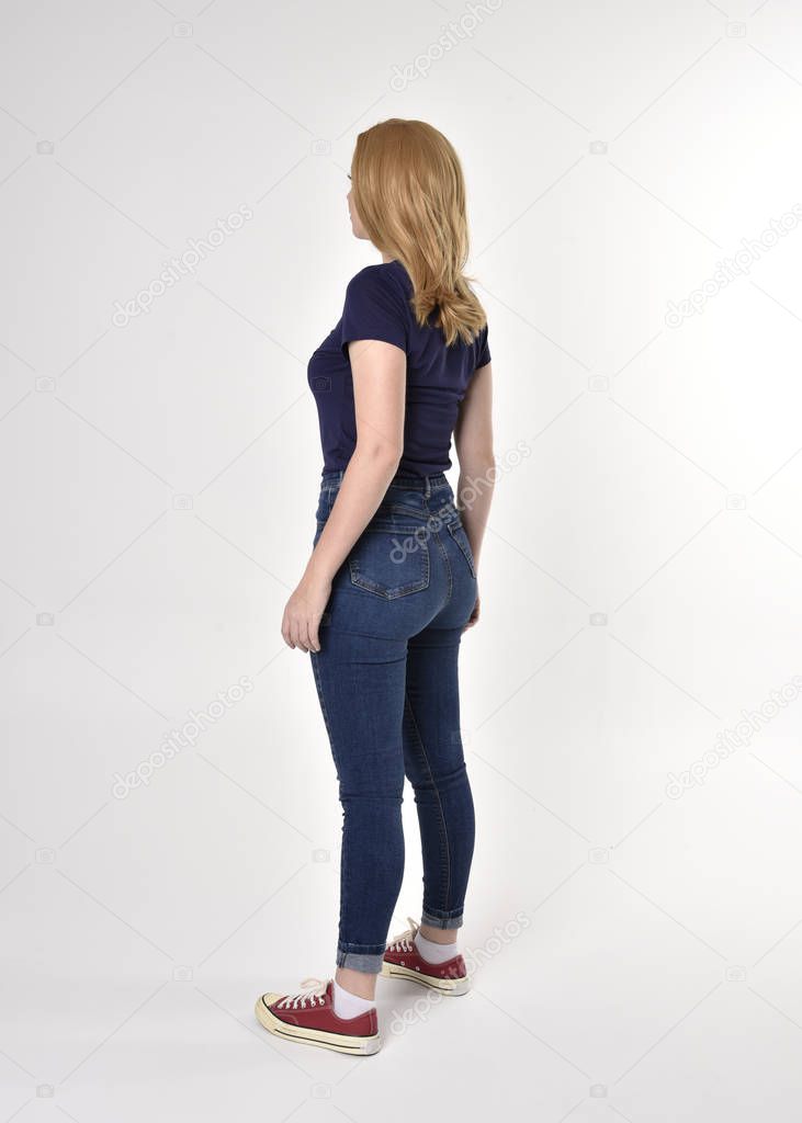 Full length portrait of a pretty blonde girl wearing casual blue shirt, denim jeans and sneakers. Standing pose, walking away from the camera, on a studio background.