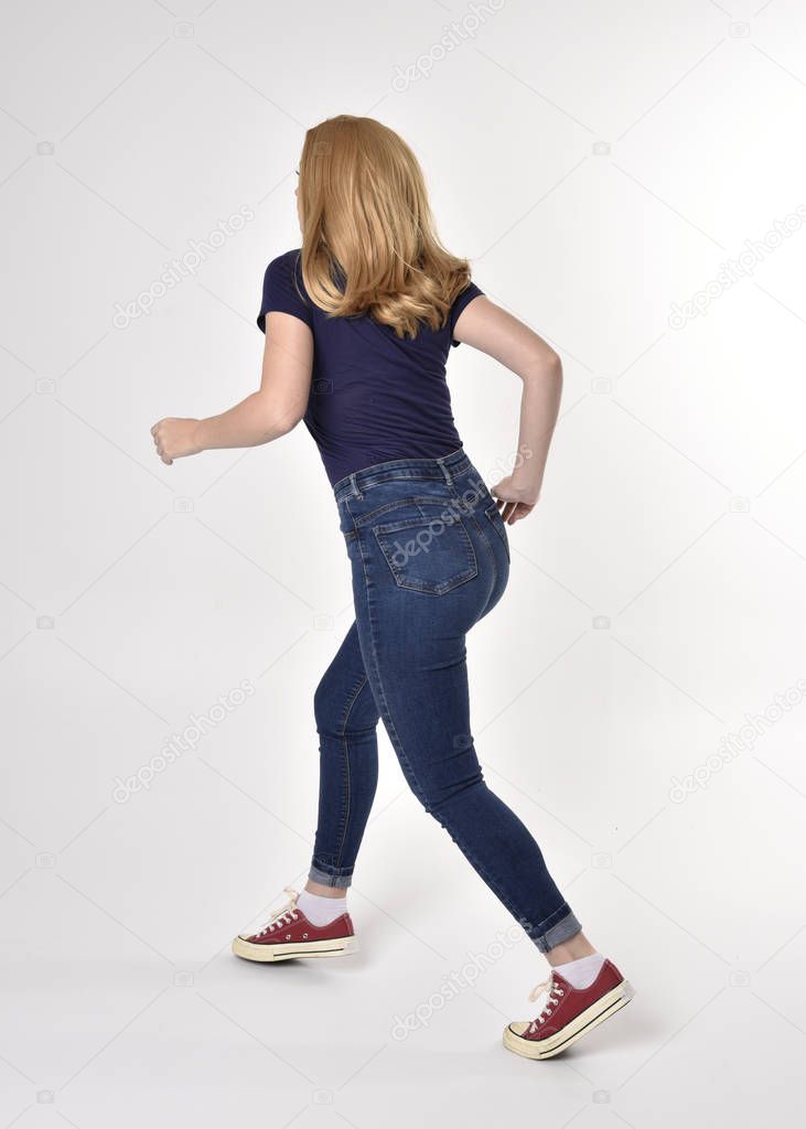 Full length portrait of a pretty blonde girl wearing casual blue shirt, denim jeans and sneakers. Standing pose, walking away from the camera, on a studio background.
