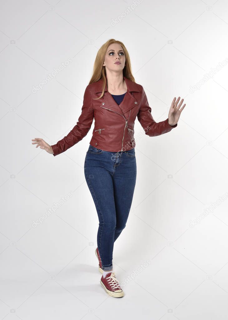Full length portrait of a pretty blonde girl wearing red leather jacket denim jeans and sneakers. Standing pose  on a studio background.