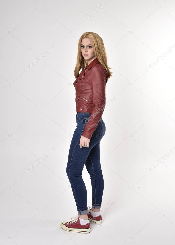 Full length portrait of a pretty blonde girl wearing red leather jacket denim jeans and sneakers. Standing pose, facing away from the camera,   on a studio background.