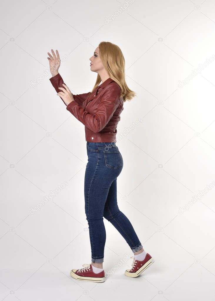Full length portrait of a pretty blonde girl wearing red leather jacket denim jeans and sneakers. Standing pose, facing away from the camera,   on a studio background.