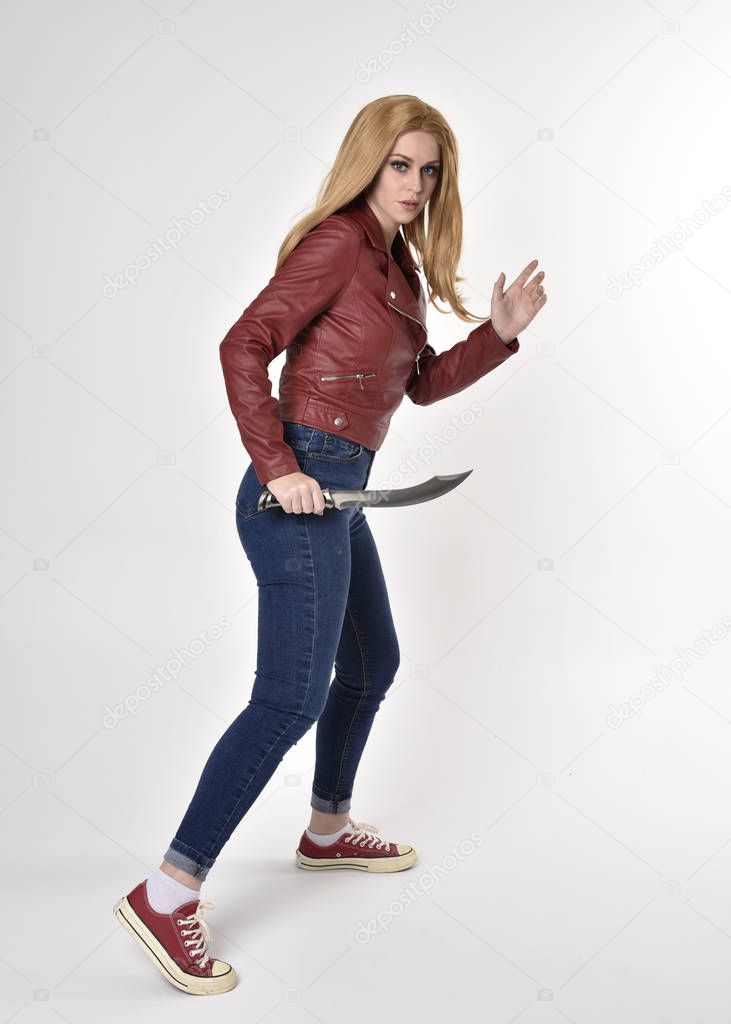 Full length portrait of a pretty blonde girl wearing red leather jacket denim jeans and sneakers. Standing pose, holding a dagger,  on a studio background.