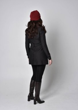 Siyah ceketli, deri çizmeli ve kırmızı bereli güzel esmer bir kızın tam boy portresi. Gri bir stüdyonun arka planında dikiliyor..