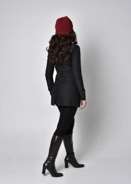 Siyah ceketli, deri çizmeli ve kırmızı bereli güzel esmer bir kızın tam boy portresi. Gri bir stüdyonun arka planında dikiliyor..