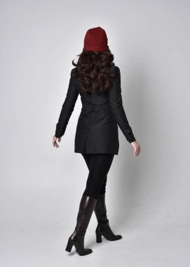 Siyah ceketli, deri çizmeli ve kırmızı bereli güzel esmer bir kızın tam boy portresi. Gri bir stüdyonun arka planında dikiliyor..