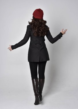 Siyah ceketli, deri çizmeli ve kırmızı bereli güzel esmer bir kızın tam boy portresi. Gri bir stüdyonun arka planında dikiliyor..