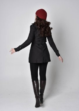 Siyah ceketli, deri çizmeli ve kırmızı bereli güzel esmer bir kızın tam boy portresi. Gri bir stüdyonun arka planında dikiliyor..