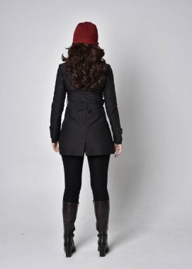 Siyah ceketli, deri çizmeli ve kırmızı bereli güzel esmer bir kızın tam boy portresi. Gri bir stüdyonun arka planında dikiliyor..