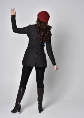 Siyah ceketli, deri çizmeli ve kırmızı bereli güzel esmer bir kızın tam boy portresi. Gri bir stüdyonun arka planında dikiliyor..