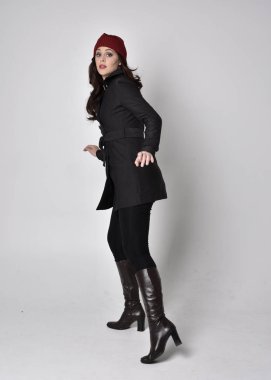 Siyah ceketli, deri çizmeli ve kırmızı bereli güzel esmer bir kızın tam boy portresi. Gri bir stüdyonun arka planında dikiliyor..