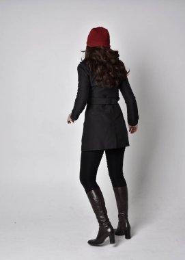 Siyah ceketli, deri çizmeli ve kırmızı bereli güzel esmer bir kızın tam boy portresi. Gri bir stüdyonun arka planında dikiliyor..