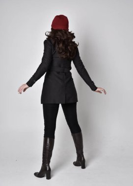 Siyah ceketli, deri çizmeli ve kırmızı bereli güzel esmer bir kızın tam boy portresi. Gri bir stüdyonun arka planında dikiliyor..