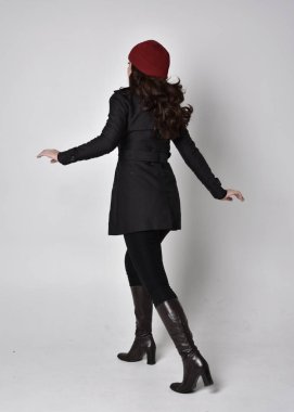 Siyah ceketli, deri çizmeli ve kırmızı bereli güzel esmer bir kızın tam boy portresi. Gri bir stüdyonun arka planında dikiliyor..