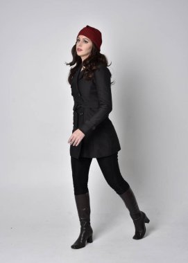 Siyah ceketli, deri çizmeli ve kırmızı bereli güzel esmer bir kızın tam boy portresi. Gri bir stüdyonun arka planında dikiliyor..