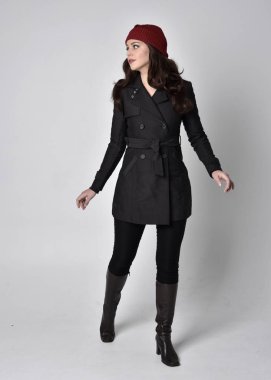 Siyah ceketli, deri çizmeli ve kırmızı bereli güzel esmer bir kızın tam boy portresi. Gri bir stüdyonun arka planında dikiliyor..