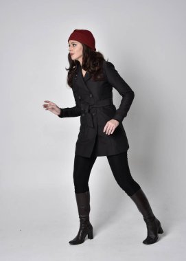 Siyah ceketli, deri çizmeli ve kırmızı bereli güzel esmer bir kızın tam boy portresi. Gri bir stüdyonun arka planında dikiliyor..