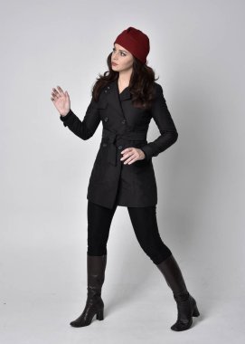 Siyah ceketli, deri çizmeli ve kırmızı bereli güzel esmer bir kızın tam boy portresi. Gri bir stüdyonun arka planında dikiliyor..