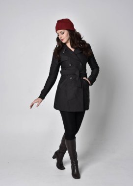 Siyah ceketli, deri çizmeli ve kırmızı bereli güzel esmer bir kızın tam boy portresi. Gri bir stüdyonun arka planında dikiliyor..