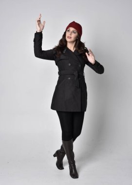 Siyah ceketli, deri çizmeli ve kırmızı bereli güzel esmer bir kızın tam boy portresi. Gri bir stüdyonun arka planında dikiliyor..