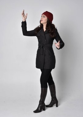 Siyah ceketli, deri çizmeli ve kırmızı bereli güzel esmer bir kızın tam boy portresi. Gri bir stüdyonun arka planında dikiliyor..