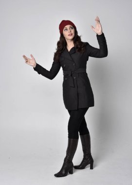 Siyah ceketli, deri çizmeli ve kırmızı bereli güzel esmer bir kızın tam boy portresi. Gri bir stüdyonun arka planında dikiliyor..