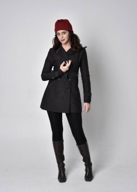 Siyah ceketli, deri çizmeli ve kırmızı bereli güzel esmer bir kızın tam boy portresi. Gri bir stüdyonun arka planında dikiliyor..