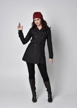 Siyah ceketli, deri çizmeli ve kırmızı bereli güzel esmer bir kızın tam boy portresi. Gri bir stüdyonun arka planında dikiliyor..