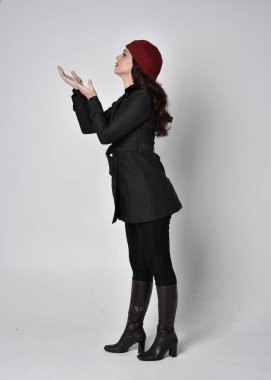 Siyah ceketli, deri çizmeli ve kırmızı bereli güzel esmer bir kızın tam boy portresi. Gri bir stüdyonun arka planında dikiliyor..